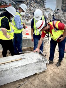 Four people in hard hats and bright yellow safety vests inspect a piece of concrete. Debris is piled up behind them.