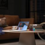 A table in the living room with a cup of tea, a laptop and a stack of books on top. Behind the table is a couch and a bike