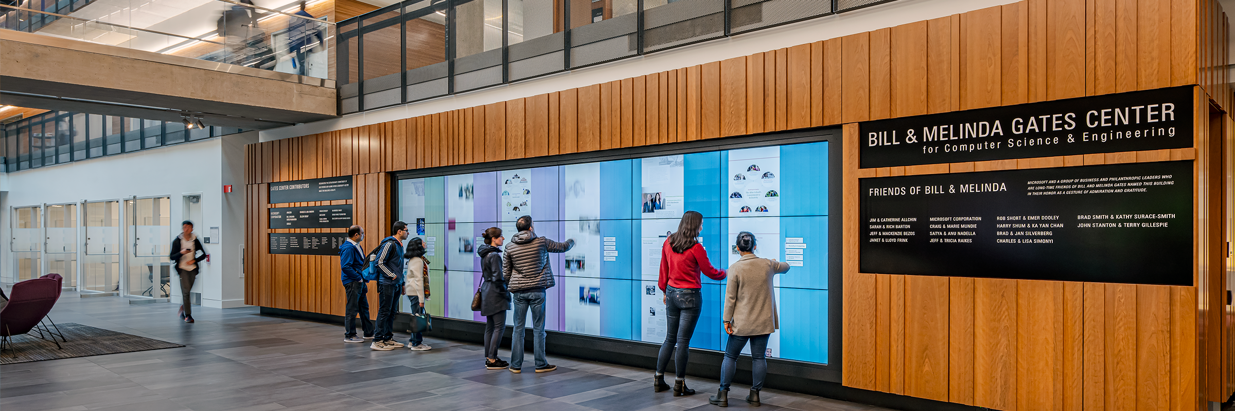 an image of the Gates Center atrium, with 7 people interacting with a large wall of touchscreen monitors next to a large sign that reads 